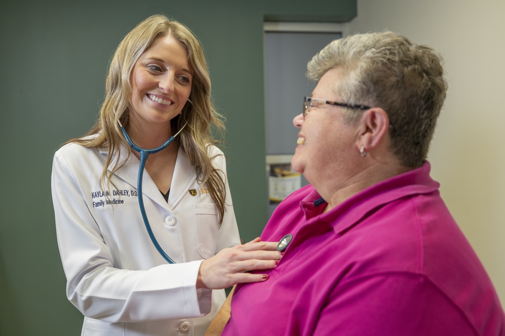 A doctor in a white coat listens to a patient's heart while smiling at one another.