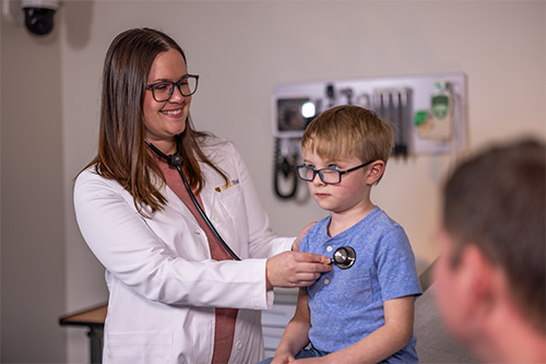 A doctor in a white coat listens to a child’s heartbeat using a stethoscope. The child is seated on an exam table. The child’s parent is partially visible in the foreground.