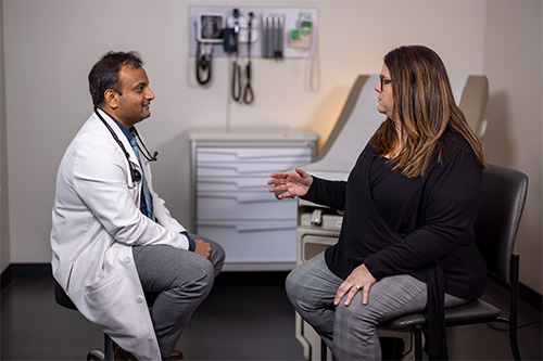 A doctor in a white coat and a patient are seated in an exam room having a conversation.