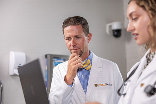 Faculty doctor in a white coat looks at a laptop screen being held by a resident in a white coat in the foreground.