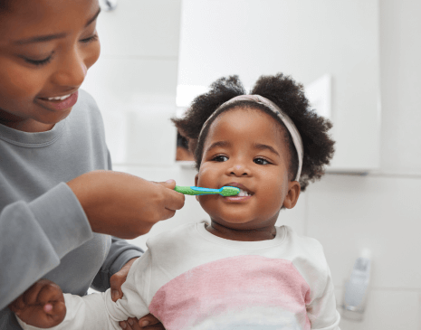 A mom brushing her daughter's teeth as she looks in the mirror.